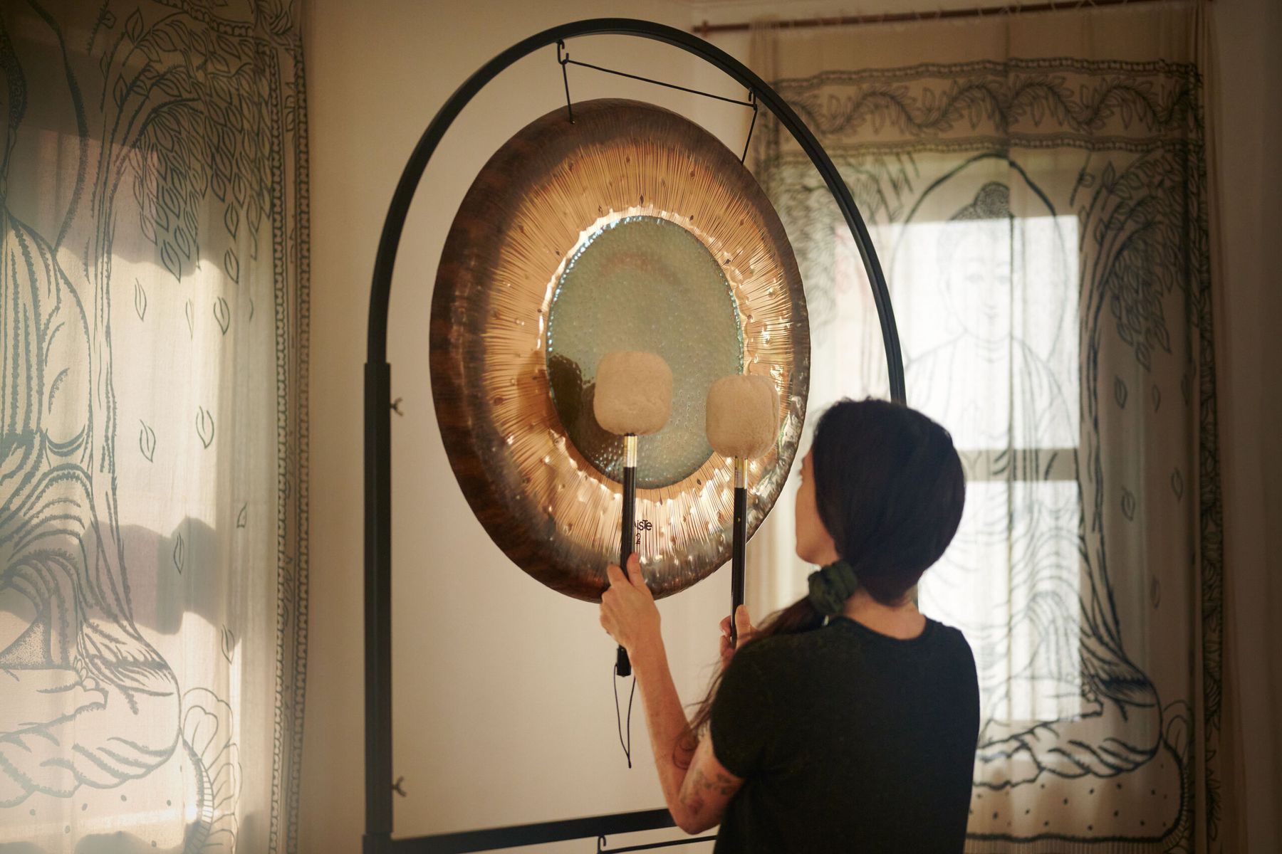 A player strikes a gong with two felt mallets beside a sunlit window