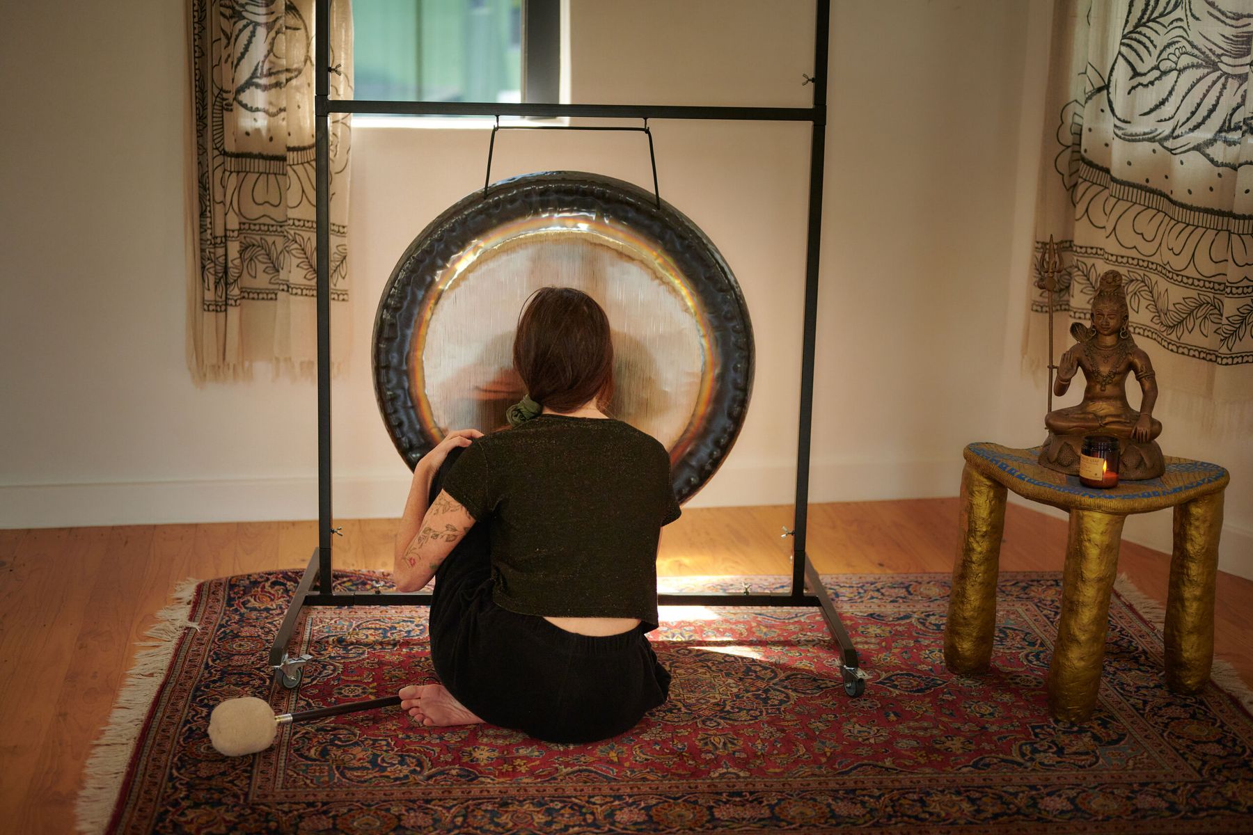 A player sits on a rug before a large gong, beside a brass statue