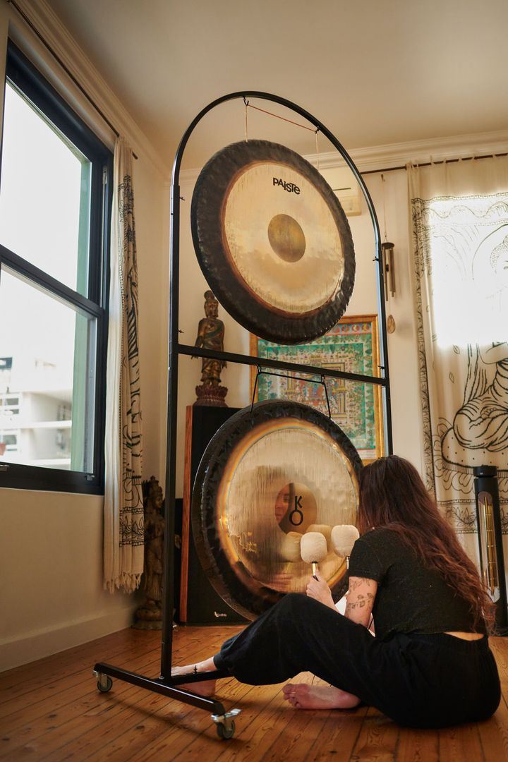 A player sits on the floor beside two gongs on a stand, by a window